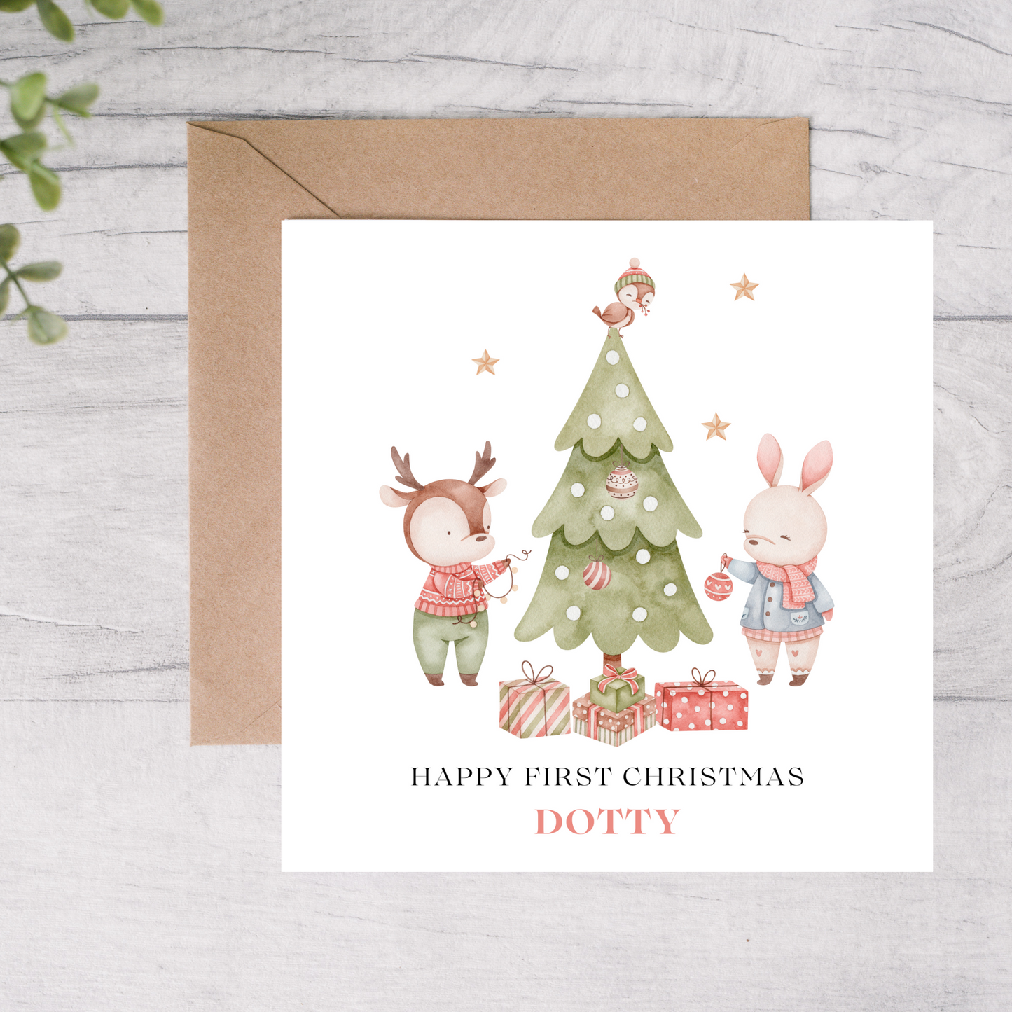 First Christmas card with a deer and rabbit decorating a tree, on a wooden surface.