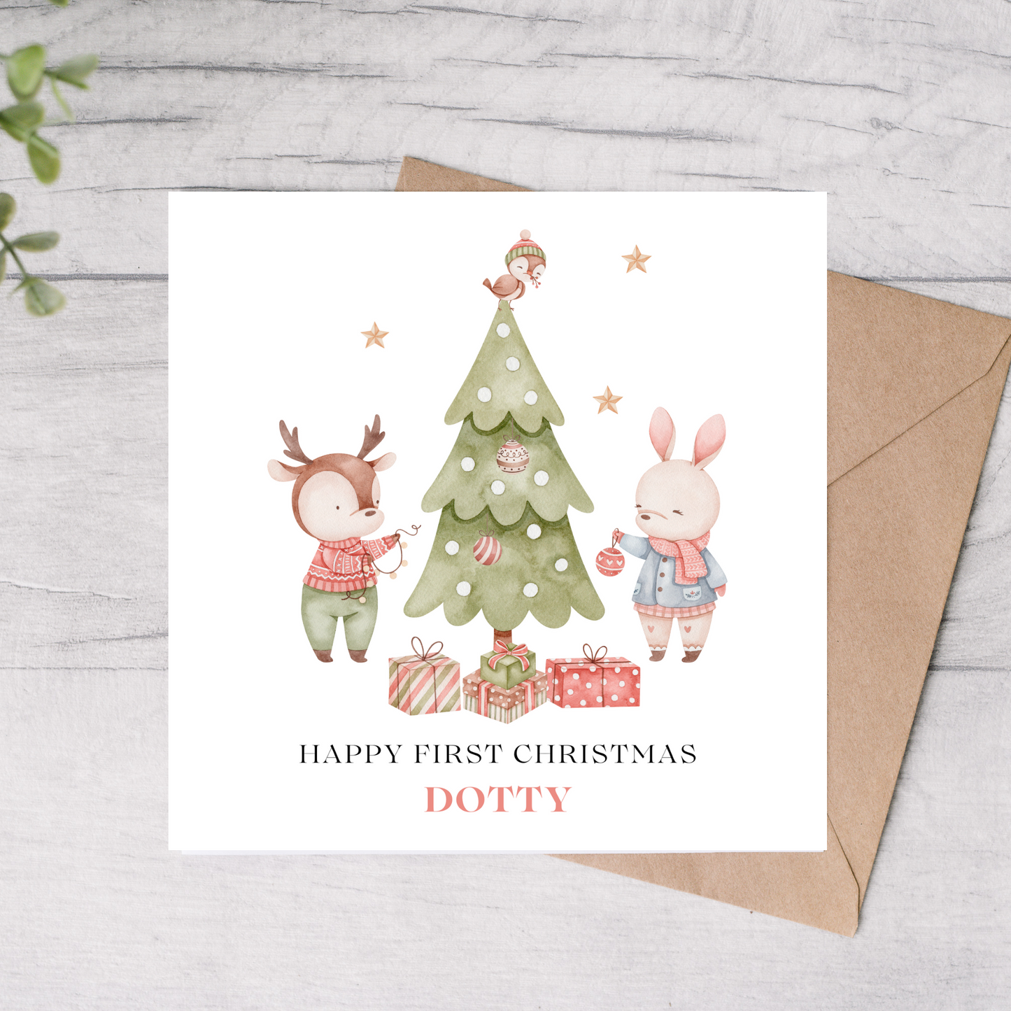 Babys 1st Christmas card with cartoon deer and rabbit by a decorated tree, on a light wooden surface.