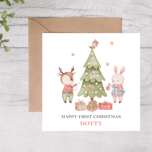 First Christmas card with a deer and rabbit decorating a tree, on a wooden surface.