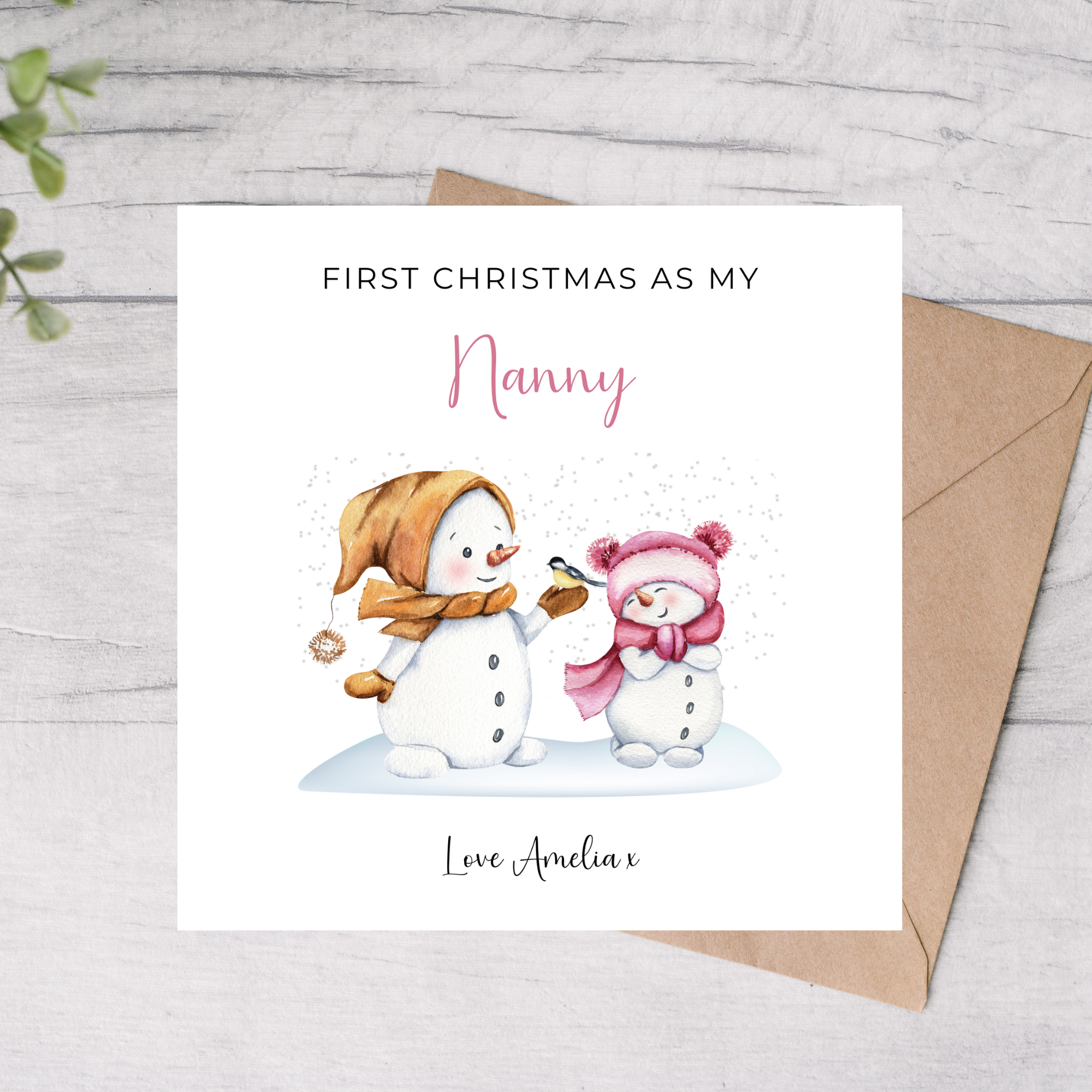 Greeting card with two snowpeople and text 'First Christmas as my Nanny' on a wooden surface.
